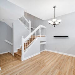 A modern, open-concept living space featuring light wooden flooring, a white staircase with wooden steps and white railings. The room has light gray walls, a white chandelier, and a small black floating shelf. The area is well-lit with natural light.