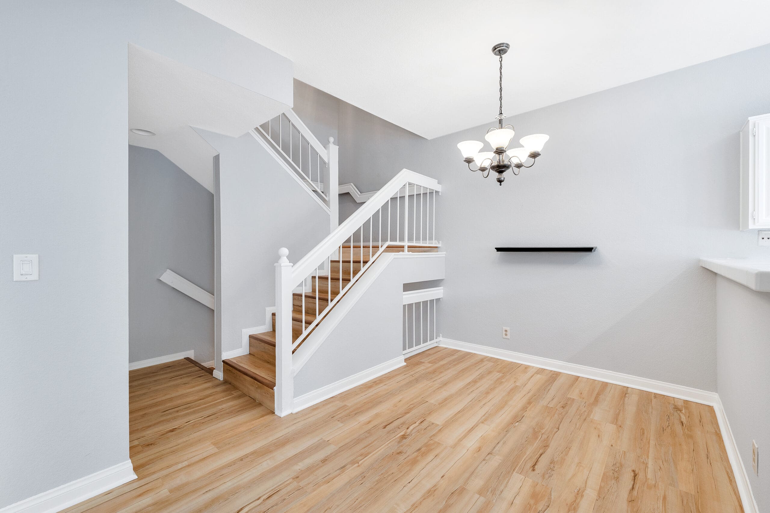 A modern, open-concept living space featuring light wooden flooring, a white staircase with wooden steps and white railings. The room has light gray walls, a white chandelier, and a small black floating shelf. The area is well-lit with natural light.