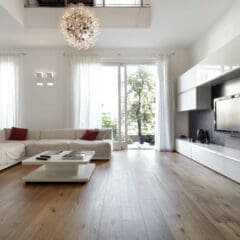 Modern living room with a light, neutral color palette. Features a large beige sectional sofa with red throw pillows, a white coffee table, a wall-mounted TV, and floor-to-ceiling windows with sheer curtains. A stylish chandelier hangs from the ceiling.