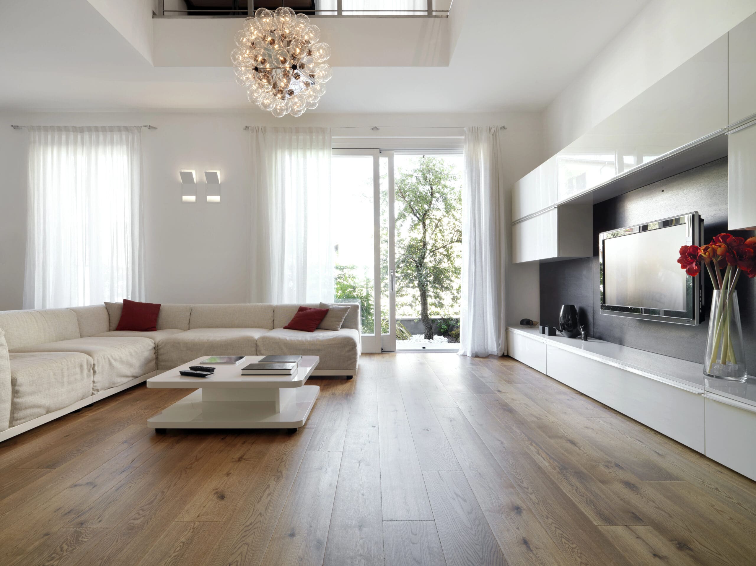 Modern living room with a light, neutral color palette. Features a large beige sectional sofa with red throw pillows, a white coffee table, a wall-mounted TV, and floor-to-ceiling windows with sheer curtains. A stylish chandelier hangs from the ceiling.