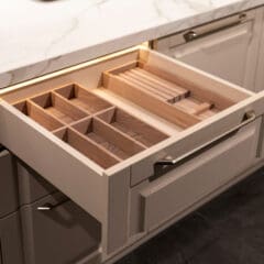 An open kitchen drawer with wooden organizers for cutlery and knives, set in a white cabinet below a white marble countertop. The drawer is empty, showing separate compartments for utensils.
