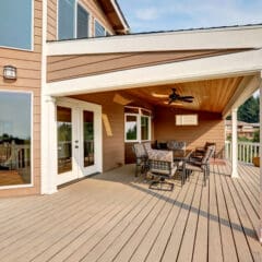 Spacious wooden deck with outdoor dining table and chairs under a covered patio attached to a tan house, surrounded by a white railing and trees in the background.