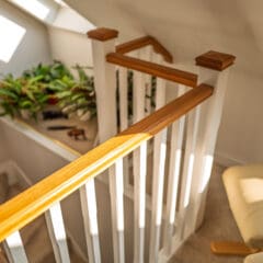 Sunlight streams onto a wooden handrail and white banister at the top of a carpeted staircase, with green potted plants on the landing and a cream-colored chair nearby.