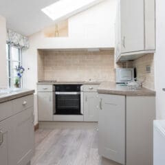 Modern kitchen with light beige cabinets, wood-style flooring, tiled backsplash, electric stove, oven, microwave, and a skylight above. A window lets in natural light, and there are flowers in a vase by the sink.