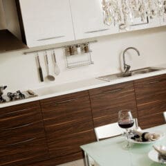 Modern kitchen with dark wood cabinets, a stovetop with a pot, utensils hanging on the wall, a sink, and spice jars. In the foreground, a glass table is set with two glasses of red wine and folded napkins.