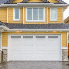 A modern yellow house with white trim featuring a large white double garage door, stone accents at the base, and a paved driveway in front. A wooden fence is visible to the right.