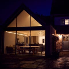 A modern house with large glass windows is warmly lit inside at night, showing a dining table and kitchen. The exterior patio and a side entrance door are dimly illuminated.