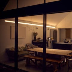 A modern dining room viewed through a large glass window at night, featuring a wooden table with bench seating, a kitchen island, and minimalist decor, including a star ornament and antlers on the wall.