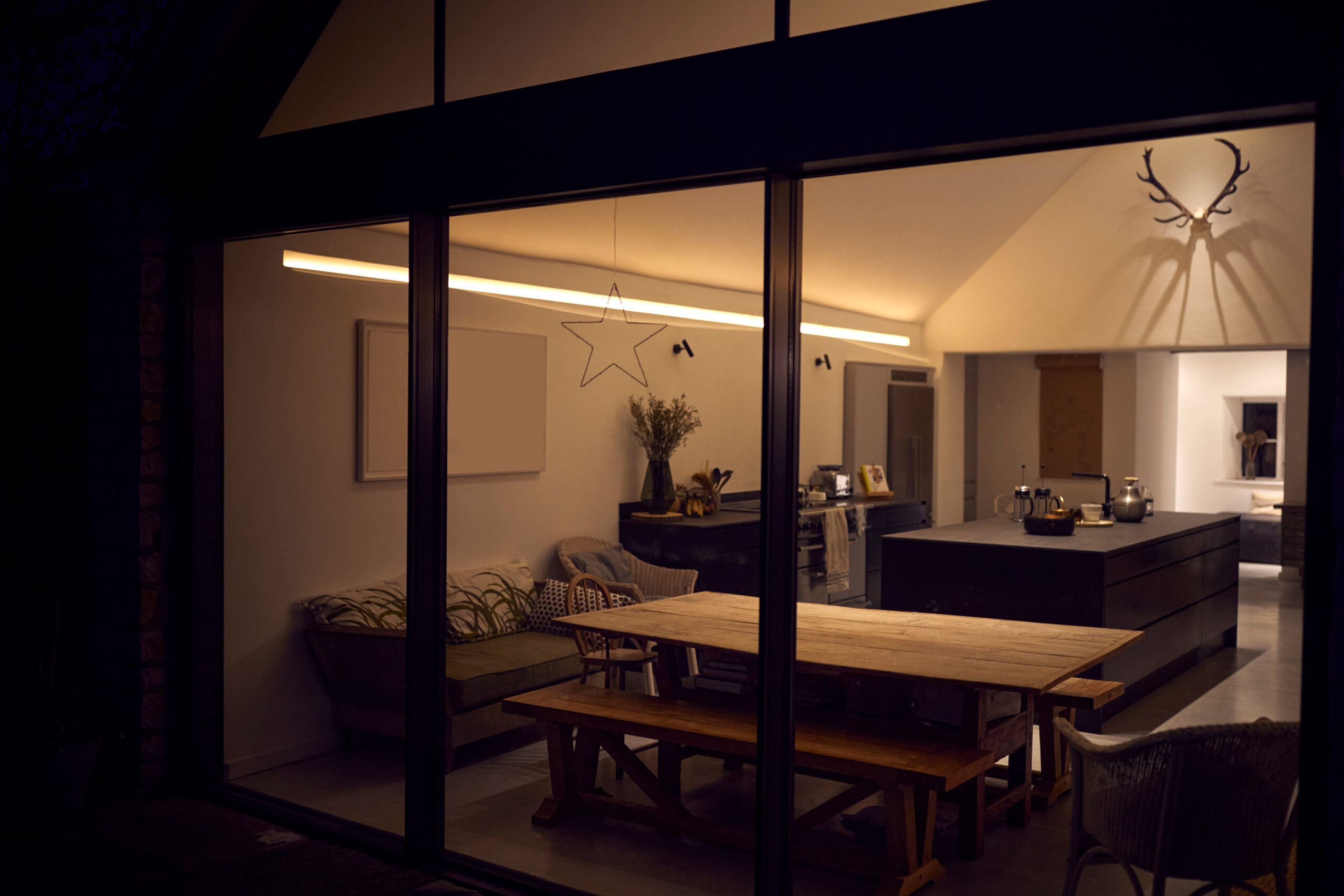 A modern dining room viewed through a large glass window at night, featuring a wooden table with bench seating, a kitchen island, and minimalist decor, including a star ornament and antlers on the wall.