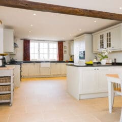 Spacious, bright kitchen with cream cabinets, a central island, wooden beams, light tiled floor, dining table with chairs, white orchid, and large window with red curtains letting in natural light.