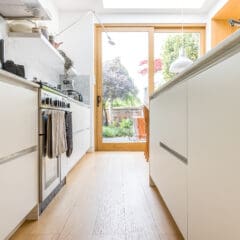 A modern, bright kitchen with white cabinets and wood flooring, featuring an oven with towels hanging, an overhead lamp, and a glass door opening to a green garden with outdoor chairs.