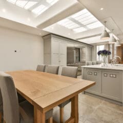 Modern kitchen and dining area with a wooden table, upholstered chairs, grey cabinetry, island with sink, pendant lights, skylights, and a vase of purple flowers on the counter. The space is bright and spacious.