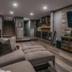 Modern basement living room with gray sectional sofa, wood flooring, wall-mounted TV, wooden cabinets, and a staircase leading upstairs. The space is warmly lit with recessed lighting and features a stone accent wall.